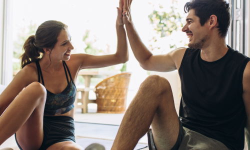 A man and a woman in workout attire giving a high five while sitting on a yoga mat in a bright, airy room with large windows.