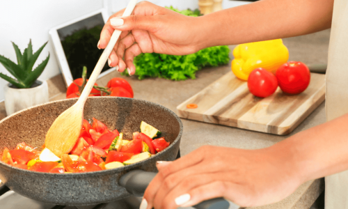 A person is stirring diced tomatoes and zucchini in a frying pan on a gas stove, with fresh vegetables on the cutting board nearby.