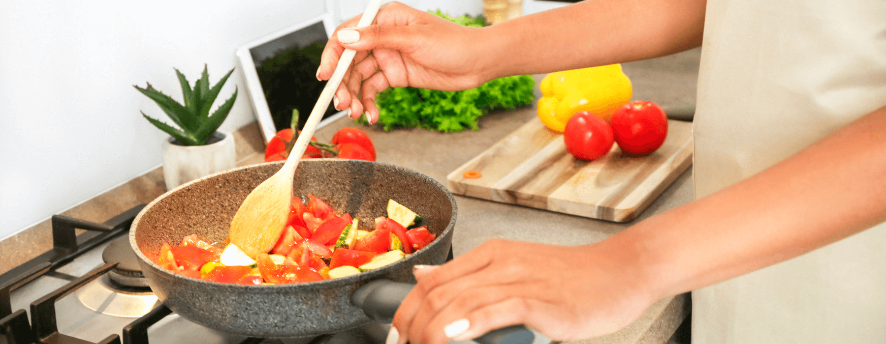 A person is stirring diced tomatoes and zucchini in a frying pan on a gas stove, with fresh vegetables on the cutting board nearby.