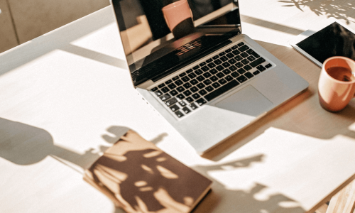 A minimalist work-from-home setup featuring a laptop, tablet, and coffee mug on a sunlit desk, with a potted plant and a book casting soft shadows, creating a cozy and productive atmosphere.