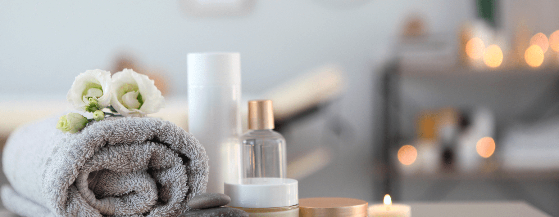 A serene spa setting featuring a rolled up grey towel adorned with white flowers, various skincare products, and softly lit candles in the background.