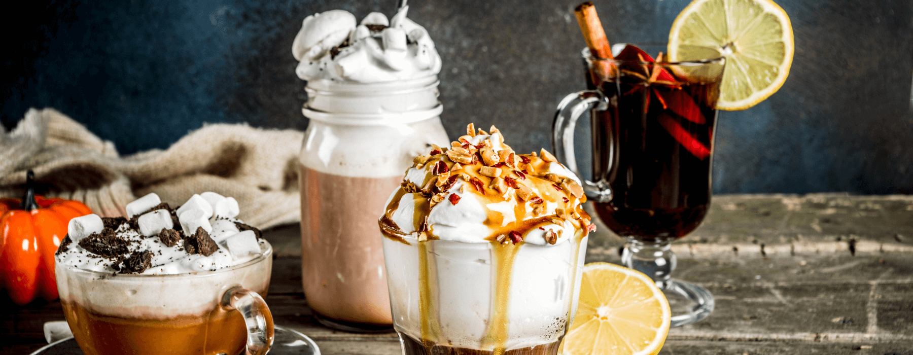 A cozy fall-themed setup showcasing a variety of festive drinks, including hot chocolate with whipped cream and marshmallows, a caramel-topped coffee with nuts, and a spiced mulled wine garnished with a lemon slice and cinnamon stick, with a small pumpkin