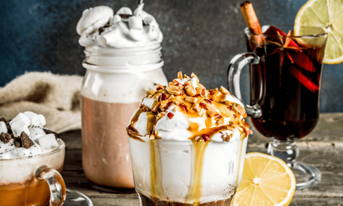 A cozy fall-themed setup showcasing a variety of festive drinks, including hot chocolate with whipped cream and marshmallows, a caramel-topped coffee with nuts, and a spiced mulled wine garnished with a lemon slice and cinnamon stick, with a small pumpkin