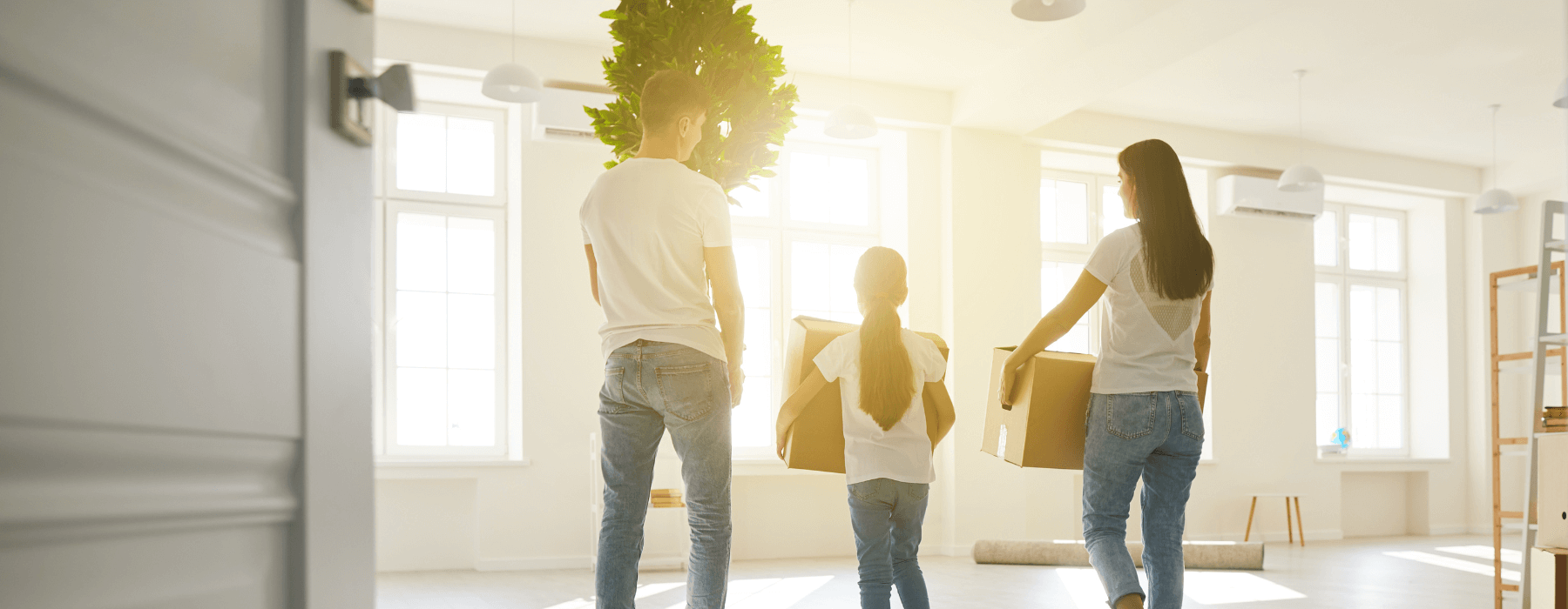 A family carrying boxes and a plant moves into a bright, sunlit room.