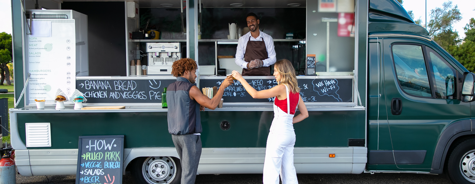 A man and a woman exchanging food at a food truck, with the vendor smiling in the background.