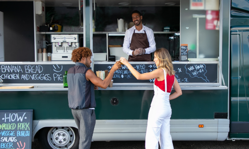 A man and a woman exchanging food at a food truck, with the vendor smiling in the background.