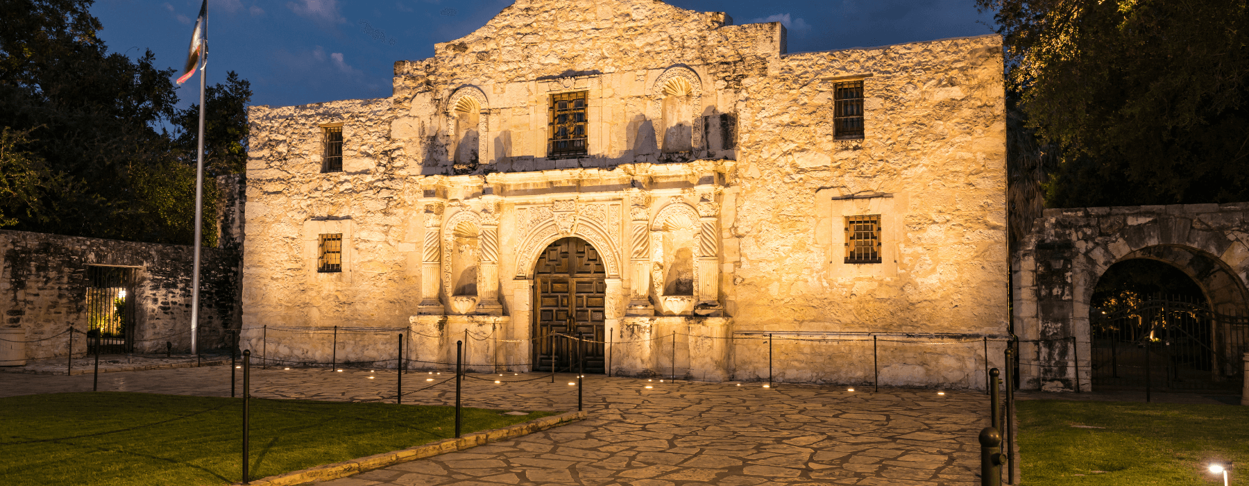 The historic Alamo at night, illuminated by warm lights, with the Texas flag flying proudly above.
