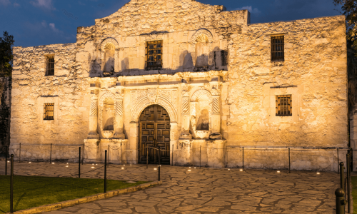 The historic Alamo at night, illuminated by warm lights, with the Texas flag flying proudly above.