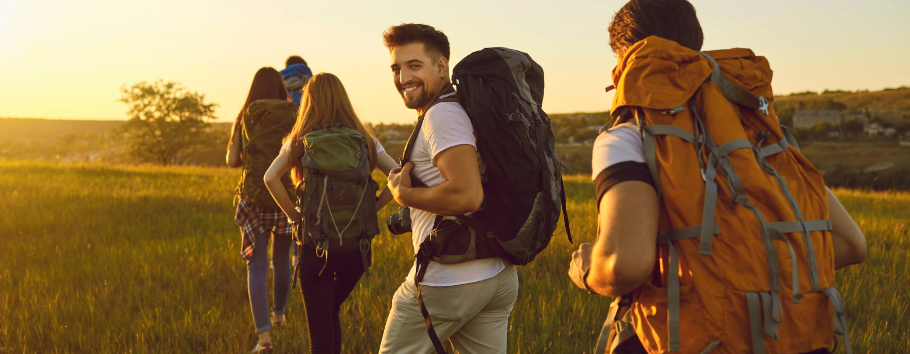 A group of friends hiking through a field at sunset, with one person smiling back at the camera