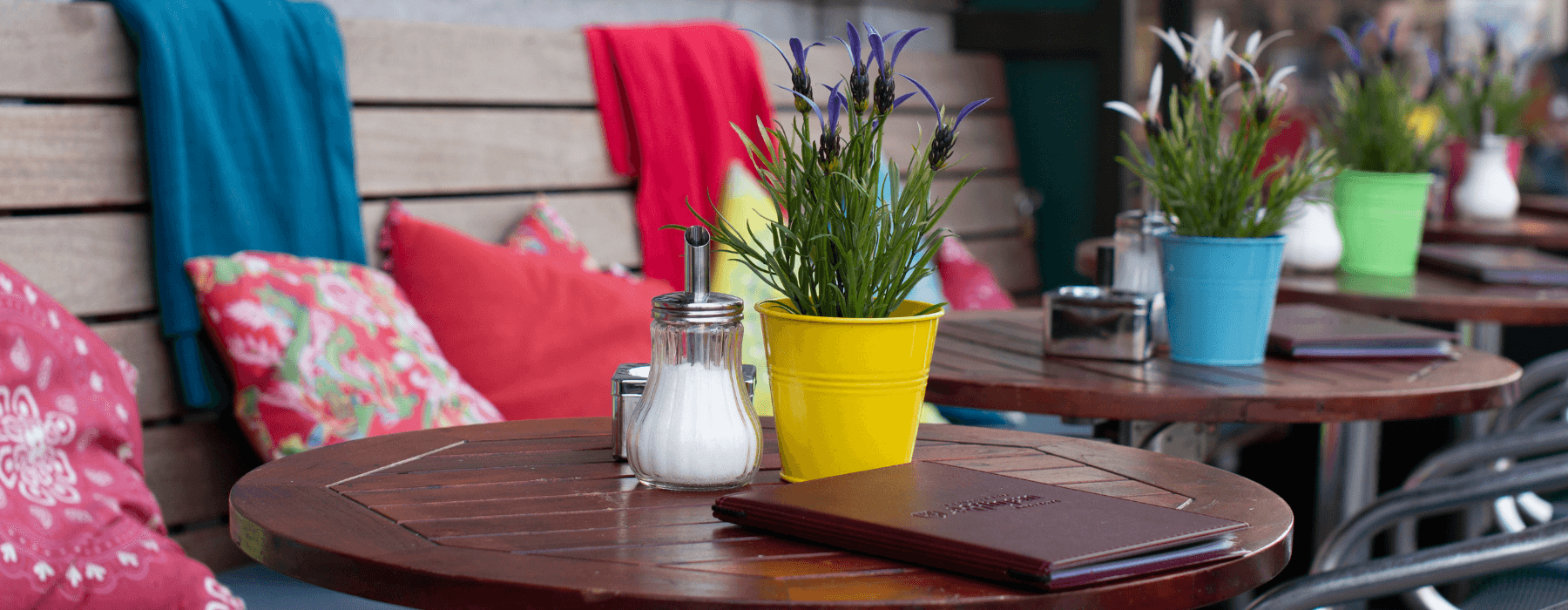 Colorful outdoor cafe tables with potted plants, cushions, and menus on a sunny day.