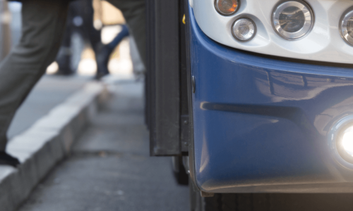 Close-up of the front section of a public bus with bright headlights, next to a curb where a passenger is stepping aboard.