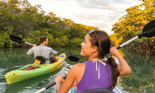 Two people kayaking through a calm, tree-lined waterway under soft, golden light.