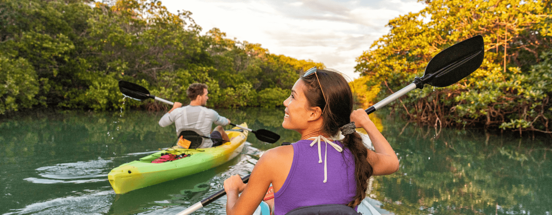 Two people kayaking through a calm, tree-lined waterway under soft, golden light.