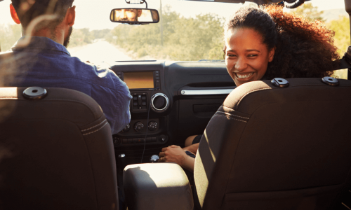 A family is on a road trip, with a woman in the passenger seat smiling back at the children in the back seat, as they drive down a sunny road.