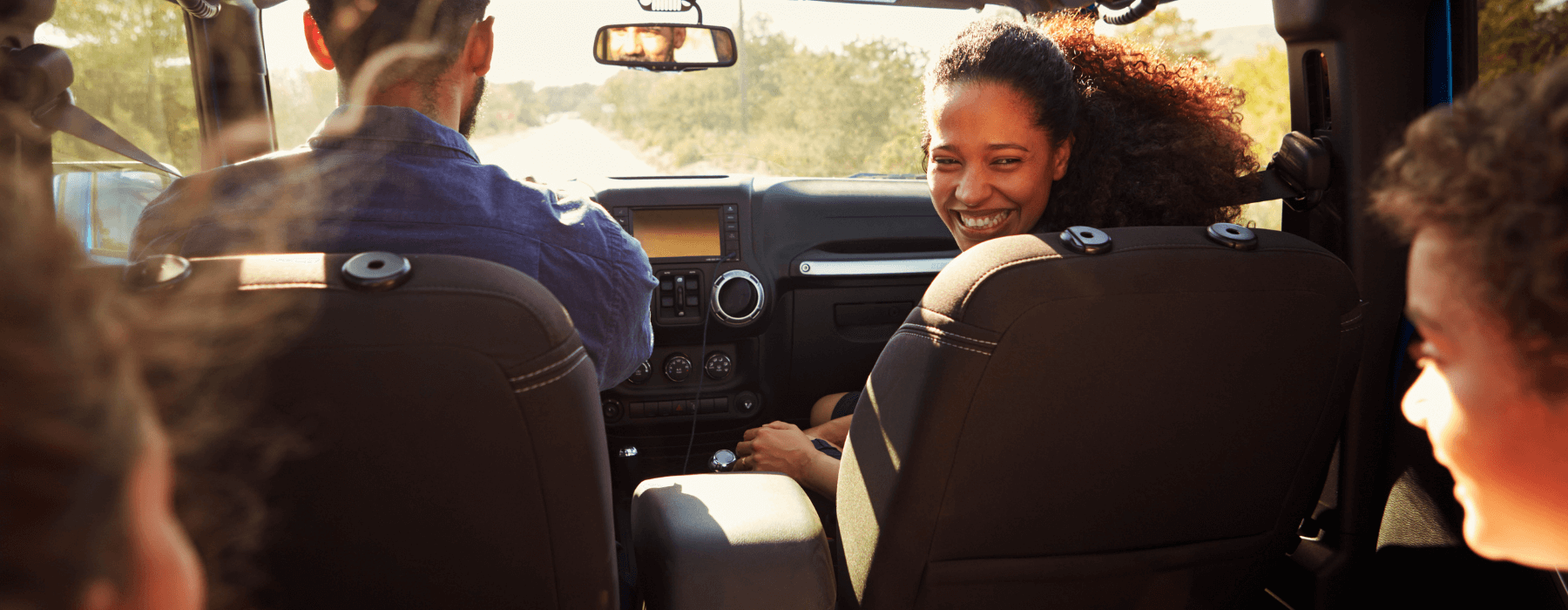 A family is on a road trip, with a woman in the passenger seat smiling back at the children in the back seat, as they drive down a sunny road.