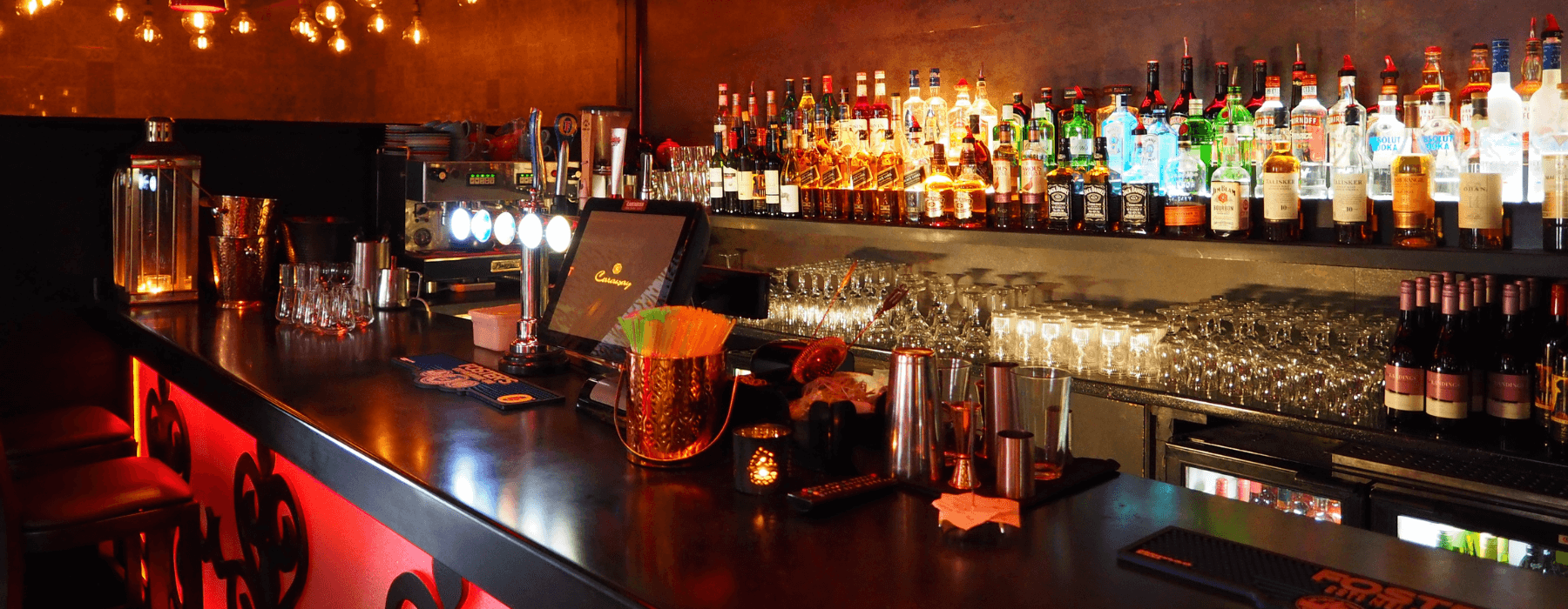 A dimly lit bar with a variety of liquor bottles, glasses, and bar tools neatly arranged.