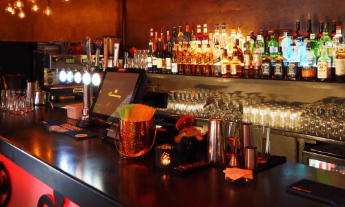 A dimly lit bar with a variety of liquor bottles, glasses, and bar tools neatly arranged.