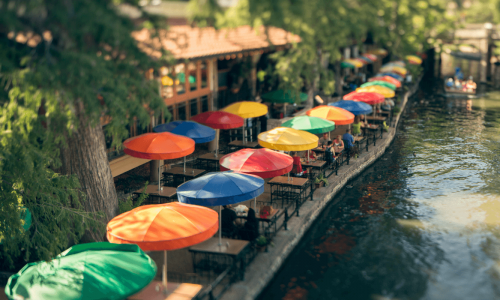 Colorful umbrellas line the outdoor seating of a riverside dining area on the San Antonio River Walk, with lush trees in the background.