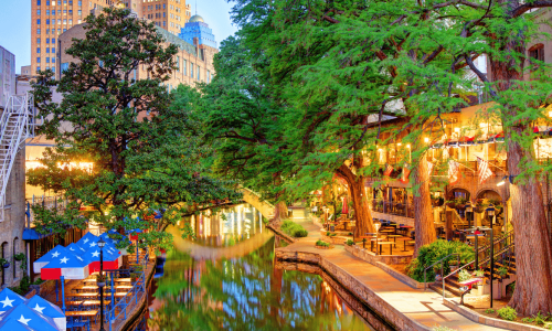 A vibrant evening scene at the San Antonio River Walk, featuring outdoor dining areas with tables and umbrellas adorned with Texas flags, surrounded by lush green trees and historic buildings reflected in the water.