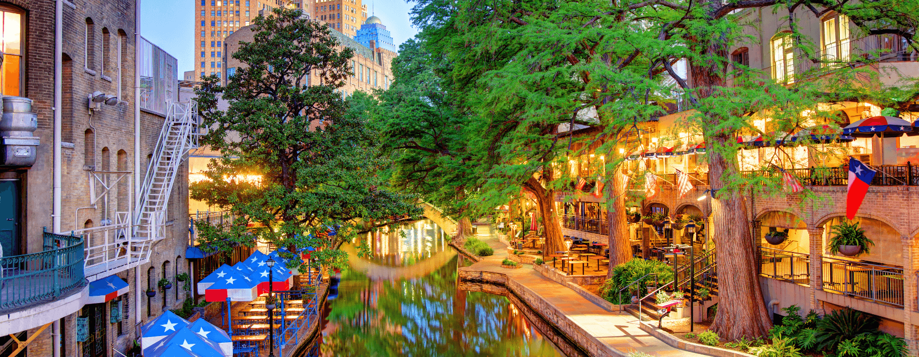 A vibrant evening scene at the San Antonio River Walk, featuring outdoor dining areas with tables and umbrellas adorned with Texas flags, surrounded by lush green trees and historic buildings reflected in the water.