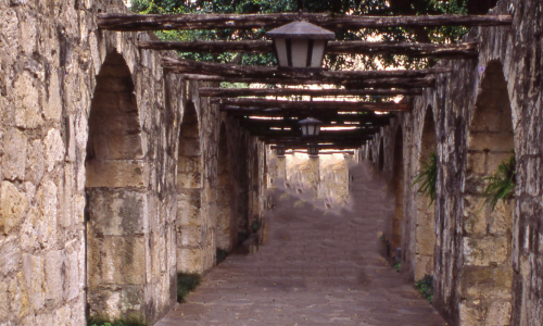 A pergola with a weathered wooden lattice roof, supported by stone pillars, over a walkway lined with ferns and street lamps.
