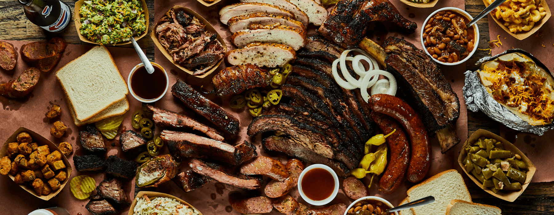 A diverse spread of barbecue dishes including smoked meats, sausages, ribs, sides like coleslaw, beans, baked potatoes, and sliced bread, arranged on a brown paper-covered table.