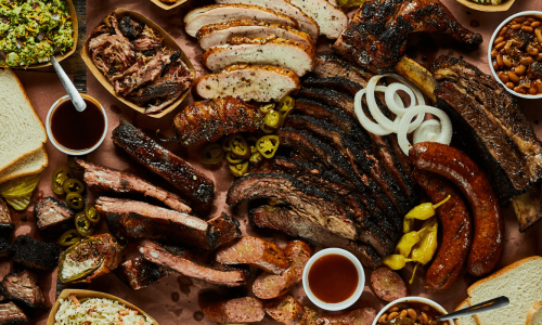 A diverse spread of barbecue dishes including smoked meats, sausages, ribs, sides like coleslaw, beans, baked potatoes, and sliced bread, arranged on a brown paper-covered table.