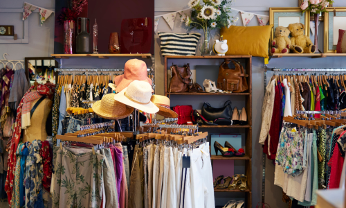 An eclectic boutique filled with racks of colorful clothing, hats, accessories, handbags, and decorative items, creating a cozy and charming shopping atmosphere.