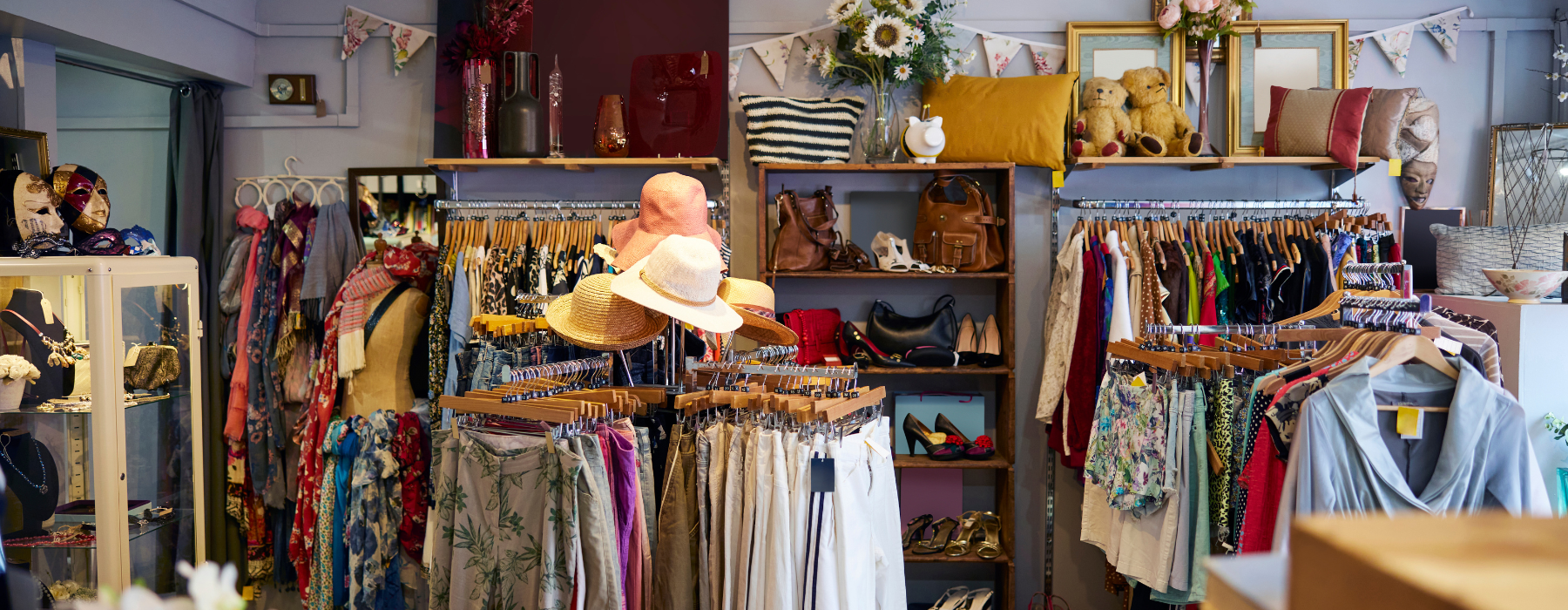 An eclectic boutique filled with racks of colorful clothing, hats, accessories, handbags, and decorative items, creating a cozy and charming shopping atmosphere.