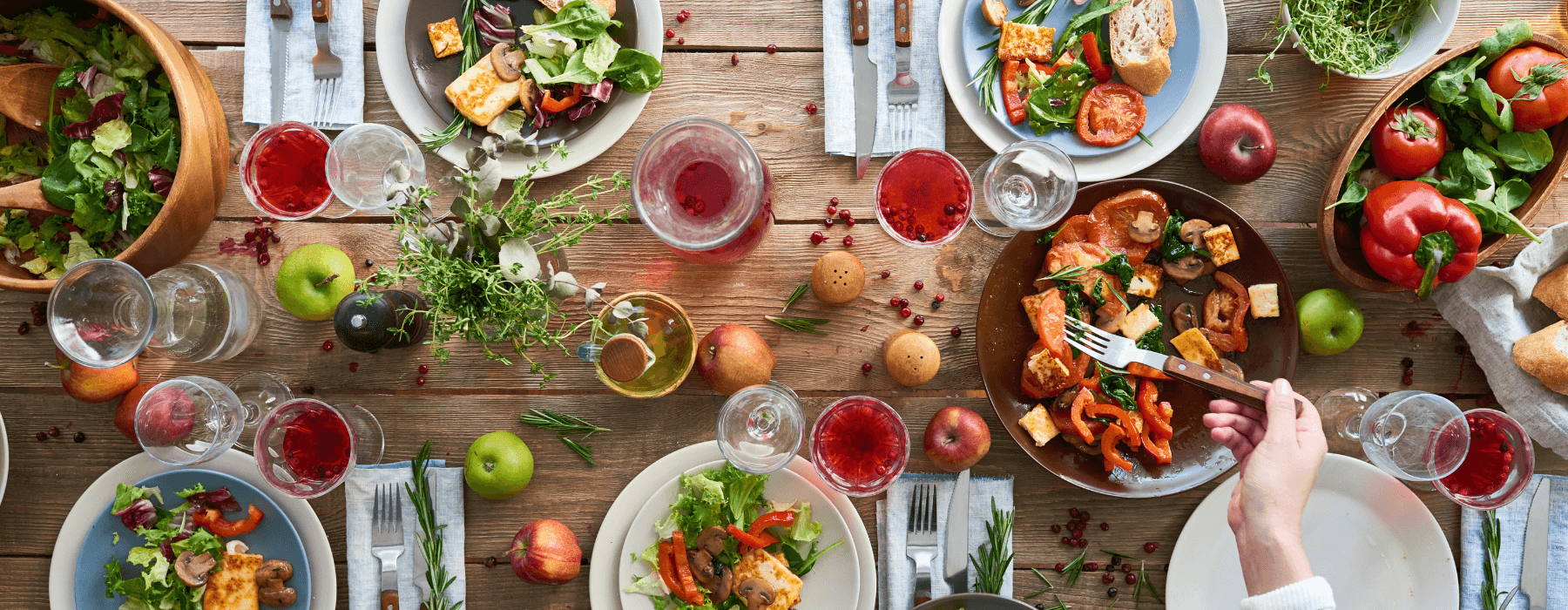 A top-down view of a rustic dining table set with fresh salads, roasted vegetables, glasses of red wine, bread, apples, and herbs, creating a vibrant and inviting meal scene.