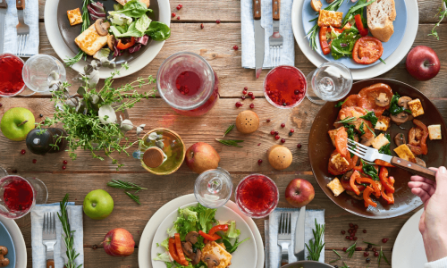 A top-down view of a rustic dining table set with fresh salads, roasted vegetables, glasses of red wine, bread, apples, and herbs, creating a vibrant and inviting meal scene.