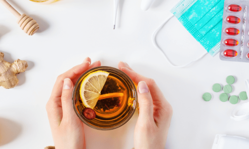 A flat lay of hands holding a mug of tea, surrounded by natural remedies like ginger, lemon, and garlic.